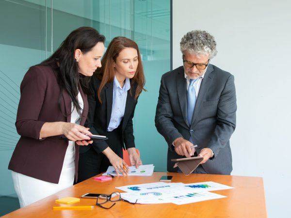 Content businesspeople watching data on tablet screen Content businesspeople watching data on tablet screen. Successful partners standing near table with statistics documents, working and watching reports in office room. Business and partnership concept